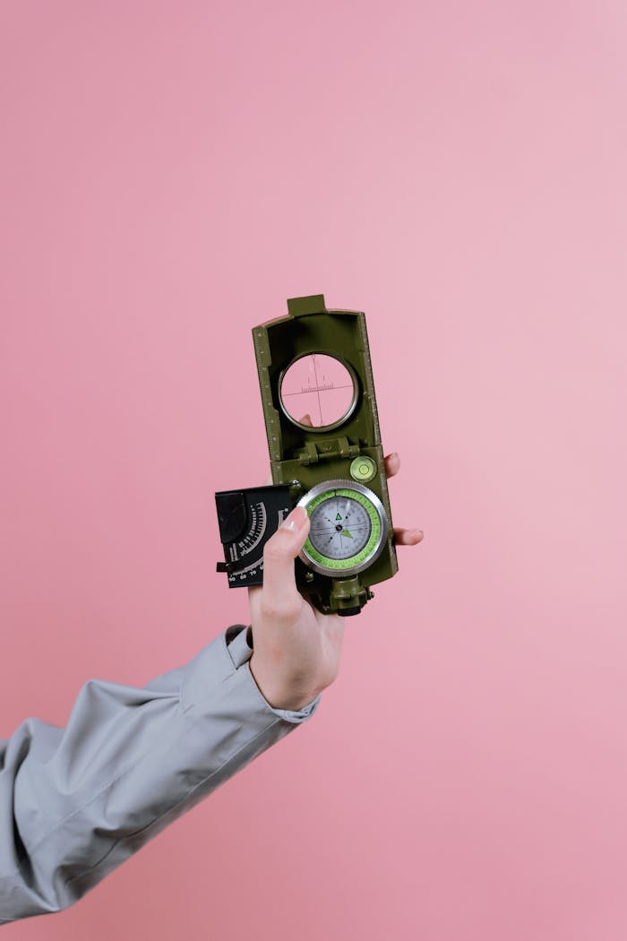 A hand holding a military compass against a pastel pink background, showcasing navigation tools.